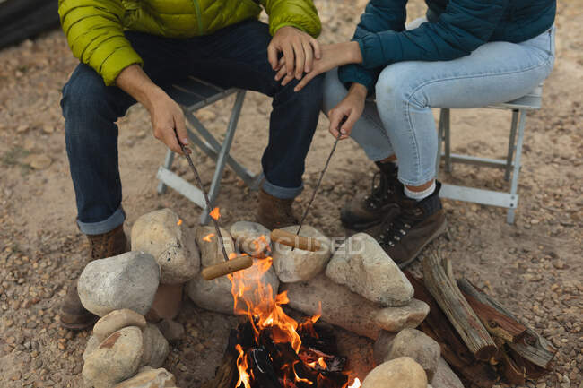 Vista frontal de ángulo alto de la pareja que se divierte en un viaje a las montañas, sentado junto a una fogata, cocinando salchichas en los palos, tomados de la mano - foto de stock