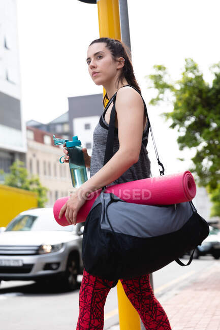 Vista lateral close-up de uma mulher caucasiana apto em seu caminho para o treinamento de fitness em um dia nublado, esperando para atravessar uma estrada, carregando um saco de esportes e um tapete de ioga, segurando uma garrafa de água — Fotografia de Stock