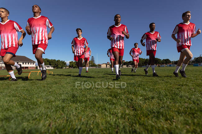 Vista frontal de ángulo bajo de un equipo masculino multiétnico adolescente de jugadores de rugby que usan su tira de equipo, corriendo juntos en el campo de juego. - foto de stock