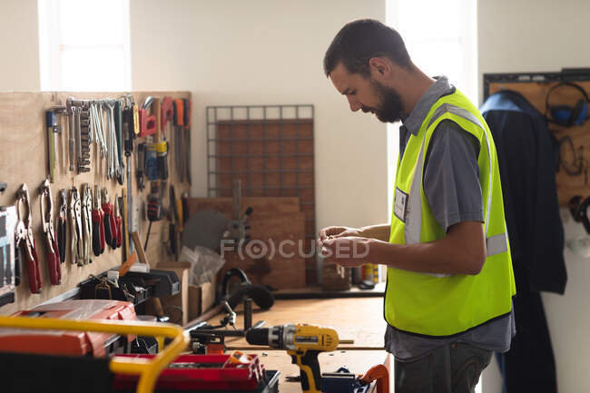 Trabalhador do sexo masculino, caucasiano, em oficina numa fábrica de cadeiras de rodas, em pé numa bancada de trabalho e a inspeccionar peças, vestindo um vestuário de trabalho — Fotografia de Stock