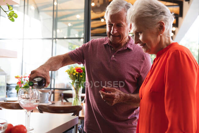 Счастливая пожилая кавказская пара дома на кухне, муж наливает им бокалы вина и оба улыбаются, дома вместе изолируя во время пандемии коронавируса — стоковое фото