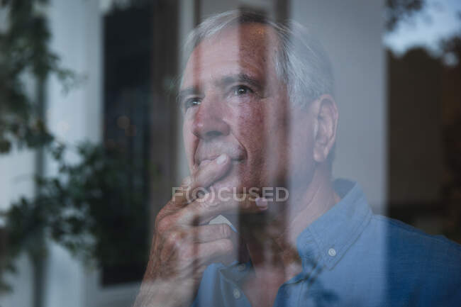 Close up de um idoso caucasiano aposentado em casa olhando para fora da janela pensando segurando seu queixo e olhando para longe, com reflexos do jardim na janela, auto-isolamento durante coronavírus covid19 pandemia — Fotografia de Stock