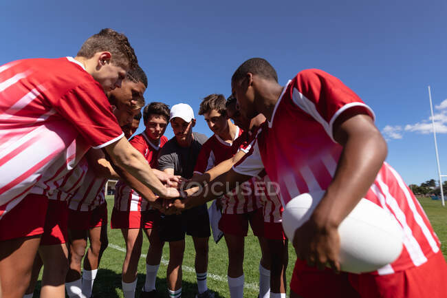Vue de face d'un entraîneur de rugby masculin caucasien empilant les mains avec une équipe masculine multi-ethnique d'adolescents de rugby portant leur bande d'équipe et se tenant debout sur le terrain de jeu. — Photo de stock
