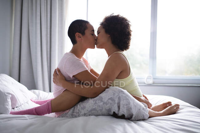 Vista lateral do casal feminino misto relaxando em casa, sentados na cama juntos pela manhã, abraçando e beijando — Fotografia de Stock