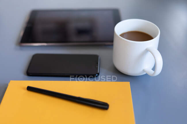 Vue rapprochée d'une tablette, d'un smartphone, d'un carnet jaune avec stylo et d'une tasse de café blanche, posé sur un bureau pendant le travail à domicile. Distance sociale et isolement personnel en quarantaine. — Photo de stock