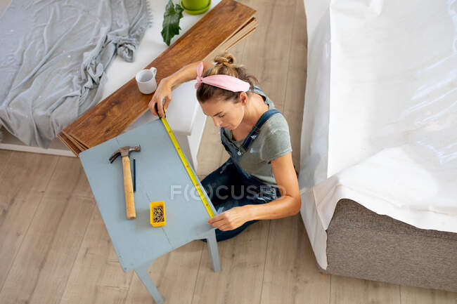 Kaukasische Frau in Blue-Jeans-Latzhose, zu Hause selbstisolierend und sozial distanziert während der Coronavirus-Epidemie 19 in Quarantäne, bastelt einen Tisch. — Stockfoto