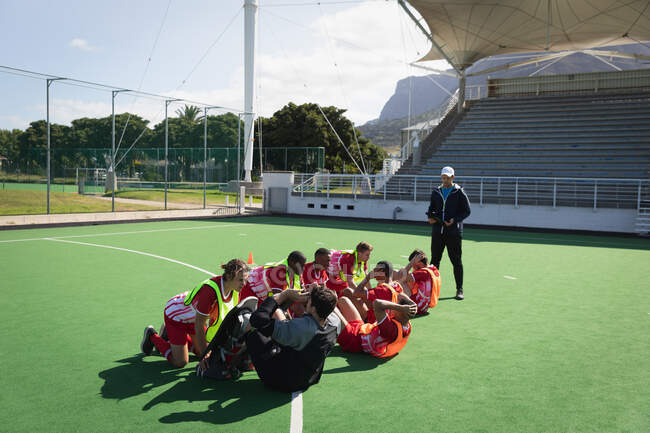 Side view of a multi-ethnic group of teenage male field hockey players and their Caucasian male field hockey coach, training before a game, working out on a field hockey pitch, doing sit ups with partners holding legs, on a sunny day — Stock Photo