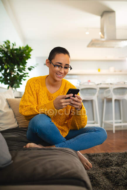 Вид женщины смешанной расы, отдыхающей дома, сидящей на диване со скрещенными ногами, использующей смартфон и улыбающейся — стоковое фото