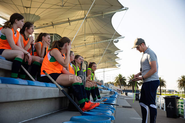 Side view of a group of female Caucasian field hockey players preparing before a game, sitting on a stand, holding hockey sticks, with their Caucasian male field hockey coach standing in front of them, talking about game plan on a sunny day — Stock Photo