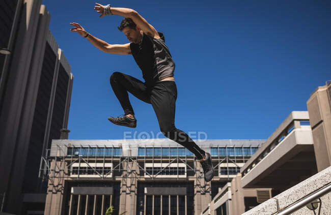 Vista lateral de um homem caucasiano praticando parkour pelo prédio em uma cidade em um dia ensolarado, pulando em corrimão de concreto. — Fotografia de Stock