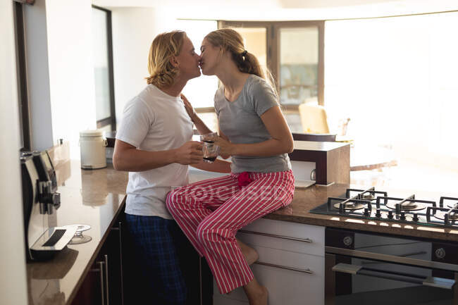 Casal caucasiano sentado em uma cozinha, bebendo café, abraçando e beijando. Distanciamento social e auto-isolamento em quarentena. — Fotografia de Stock
