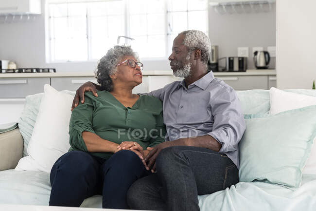 Un couple afro-américain âgé passe du temps à la maison ensemble, la distance sociale et l'isolement personnel en quarantaine pendant l'épidémie de coronavirus covid 19, assis sur un canapé, embrassant et parlant — Photo de stock