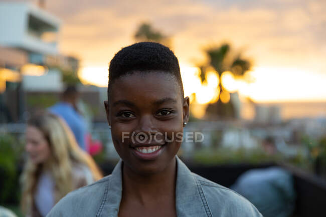 Retrato de uma mulher afro-americana pendurada num terraço com um céu de pôr-do-sol, a olhar para a câmara e a sorrir, com pessoas a falar ao fundo — Fotografia de Stock
