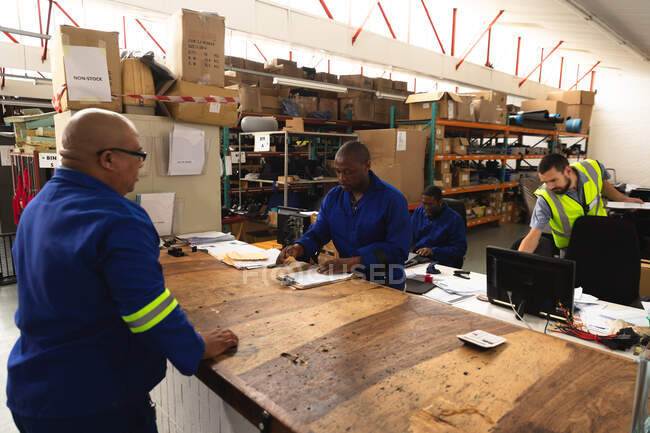 A mixed race male supervisor and an African American male worker in a storage warehouse at a factory making wheelchairs, standing and talking at a workbench — Stock Photo