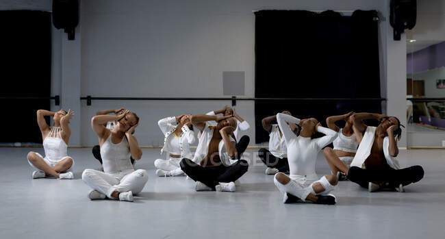 Vista frontal de un grupo multiétnico de bailarines modernos masculinos y femeninos en forma con trajes blancos que practican una rutina de baile durante una clase de baile en un estudio luminoso, sentado en el suelo. - foto de stock