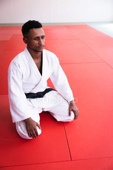 Vista frontal de ángulo alto de una judoka masculina adolescente de raza mixta que usa judogi blanco, arrodillada sobre esteras en el gimnasio antes del entrenamiento de judo. - foto de stock