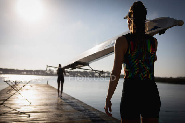 Rückansicht einer kaukasischen Ruderin, die ein Boot auf der Schulter trägt und bei Sonnenaufgang auf einem Steg am Fluss spaziert, während ihre Teamkollegin das andere Ende im Hintergrund trägt — Stockfoto