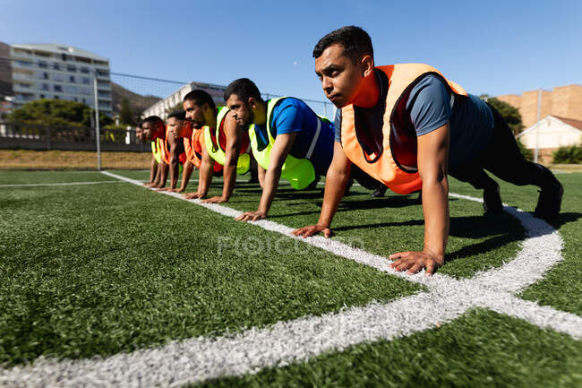 Multi gruppo etnico di cinque maschi a giocatori di calcio laterali che indossano vestiti sportivi e giubbotti di formazione in un campo sportivo al sole, riscaldandosi facendo flessioni di fila. — Foto stock