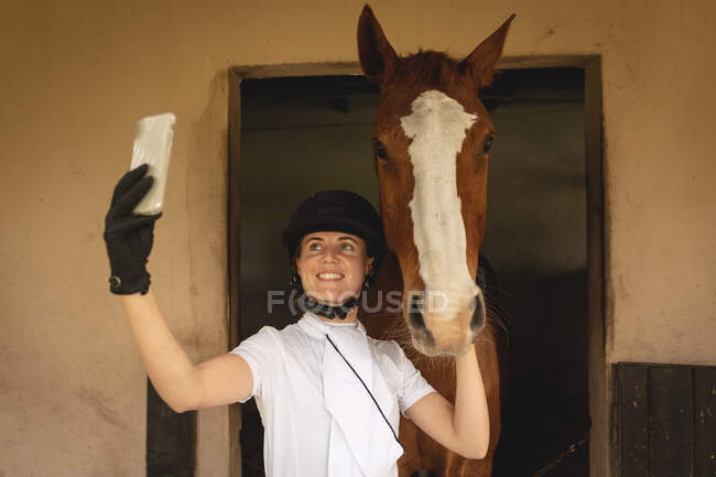 Vista frontal de cerca de una jinete caucásica casualmente vestida usando su teléfono inteligente y haciendo una selfie con su caballo castaño de pie en un establo. - foto de stock