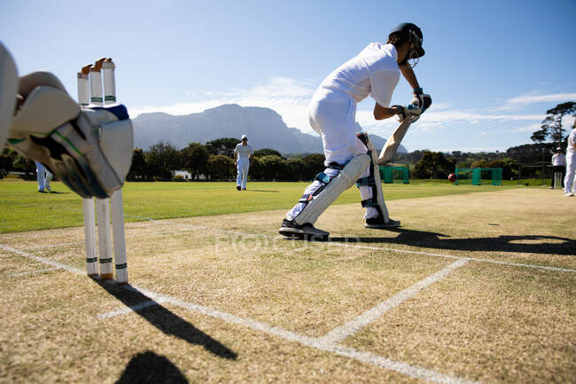 Visão traseira de um adolescente caucasiano jogador de críquete usando um capacete e segurando um taco de críquete, tentando acertar a bola em campo durante uma partida de críquete, com outros jogadores em pé no fundo. — Fotografia de Stock