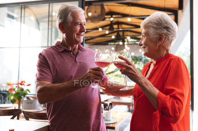 Счастливая пожилая кавказская пара дома на кухне, стоя и произнося тост с бокалами вина и улыбаясь, дома вместе изолируют во время пандемии коронавируса — стоковое фото