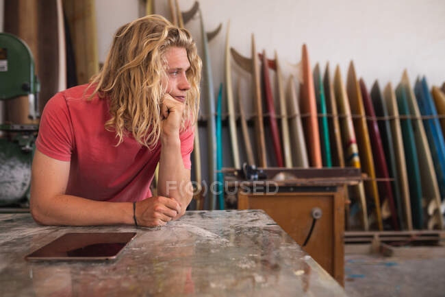 Ragionevole surfista caucasico di sesso maschile che lavora nel suo studio, appoggiandosi a un bancone e toccandosi il mento, usando un tablet, con tavole da surf in un rack sullo sfondo — Foto stock