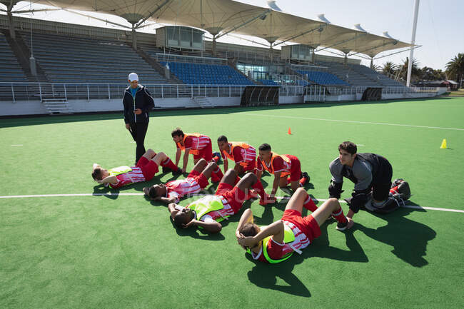 Side view of a multi-ethnic group of teenage male field hockey players and their Caucasian field hockey coach, training before a game, working out on a field hokey pitch, doing sit ups with their coach on a sunny day — Stock Photo