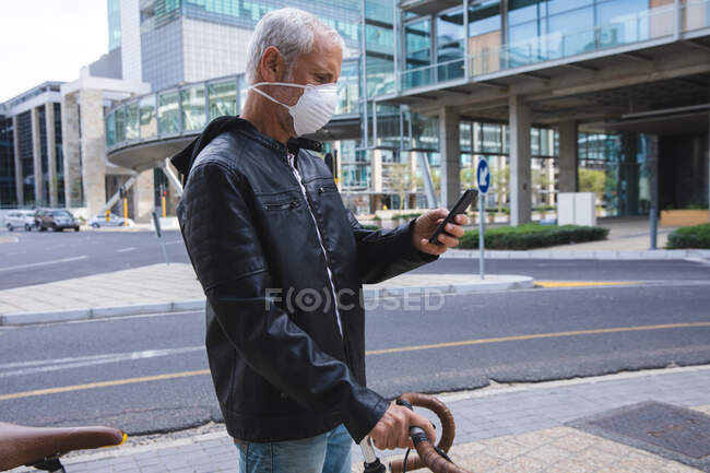 Старший кавказский мужчина днем бродит по улицам города в маске против коронавируса, ковид 19, катает велосипед и пользуется смартфоном. — стоковое фото