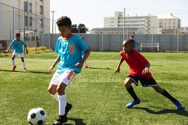 Vista frontal de una raza mixta y un niño afroamericano con sus tiras de equipo, en acción corriendo por el balón durante un partido de fútbol entre dos equipos multiétnicos de jugadores de fútbol en un campo de fútbol - foto de stock