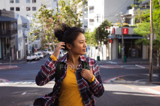 Seitenansicht einer fröhlichen Mischlingsfrau mit langen dunklen Haaren, die tagsüber in den Straßen der Stadt unterwegs ist, einen Rucksack trägt und ein kariertes Hemd trägt, das mit Gebäuden im Hintergrund lächelt. — Stockfoto
