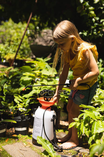 Uma menina caucasiana com longos cabelos loiros aproveitando o tempo em um jardim ensolarado, explorando, segurando um aspersor usado para plantas — Fotografia de Stock