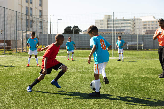 Vista lateral de dos equipos multiétnicos de jugadores de fútbol de niños que usan sus tiras de equipo, en acción durante un partido de fútbol en un campo de juego en el sol - foto de stock