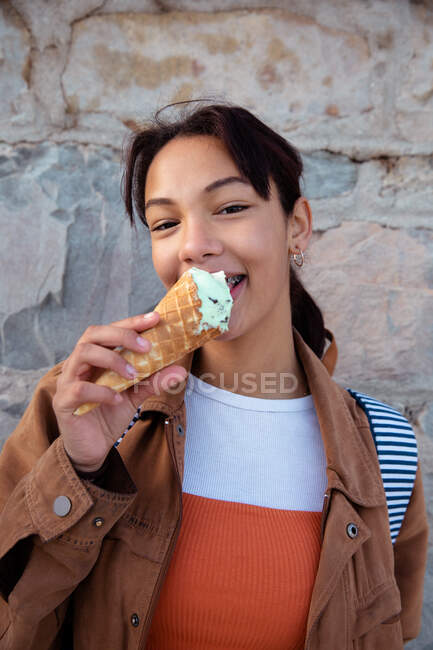 Retrato de una chica mestiza con aparatos dentales disfrutando del tiempo pasando el rato en un día soleado, parada junto a la pared, sosteniendo un helado, sonriendo directamente a la cámara. - foto de stock