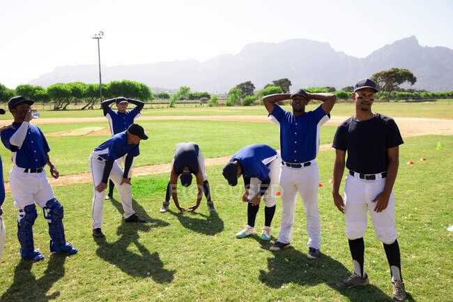 Vista lateral de un grupo multiétnico de jugadores de béisbol masculinos, entrenando antes de un juego, descansando, sentados, interactuando, en un día soleado - foto de stock