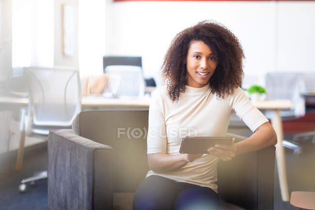 Retrato de una mujer de negocios de raza mixta con el pelo rizado, trabajando en una oficina moderna, sentada y sonriente, usando su tableta y mirando a la cámara - foto de stock