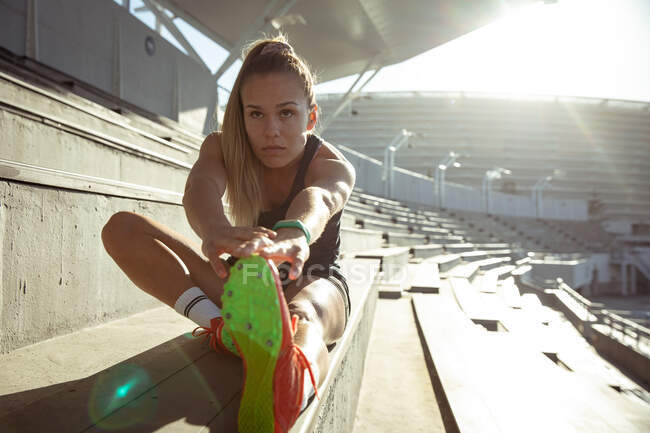Front view of a Caucasian female athlete practicing at a sports stadium, sitting in the stands and stretching, holding her foot — Stock Photo