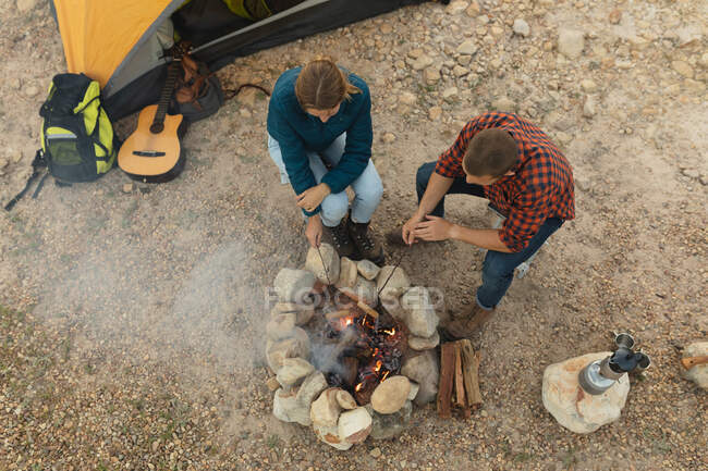 Vista de alto ângulo de um casal caucasiano se divertindo em uma viagem às montanhas, sentado perto de uma fogueira, cozinhando salsichas nas varas — Fotografia de Stock