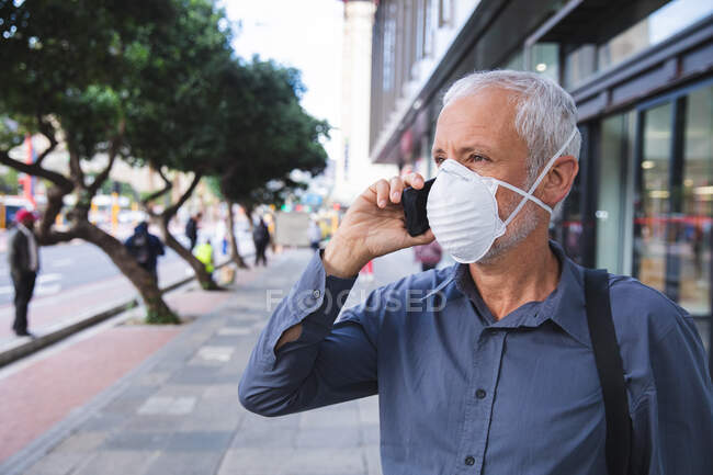 Hombre caucásico mayor por las calles de la ciudad durante el día, usando una máscara facial contra el coronavirus, covid 19 y usando un teléfono inteligente. - foto de stock