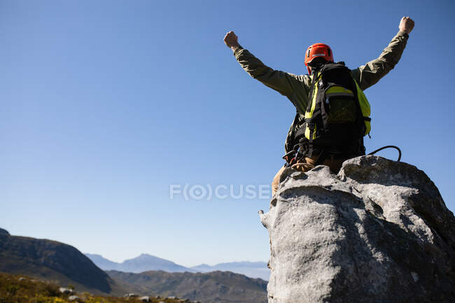 Vue arrière de l'homme caucasien profitant du temps dans la nature, portant un équipement de tyrolienne, les bras en l'air par une journée ensoleillée dans les montagnes — Photo de stock