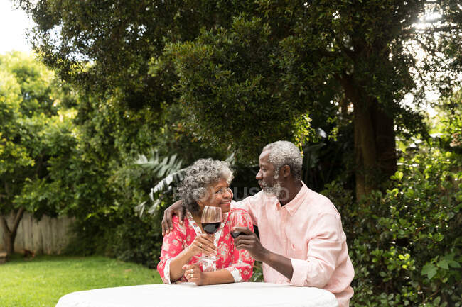 Ein älteres afroamerikanisches Paar, das während der Coronavirus-Epidemie 19 gemeinsam in seinem Garten verbringt, soziale Distanzierung und Selbstisolierung in Quarantäne, hält Gläser Rotwein und lächelt — Stockfoto