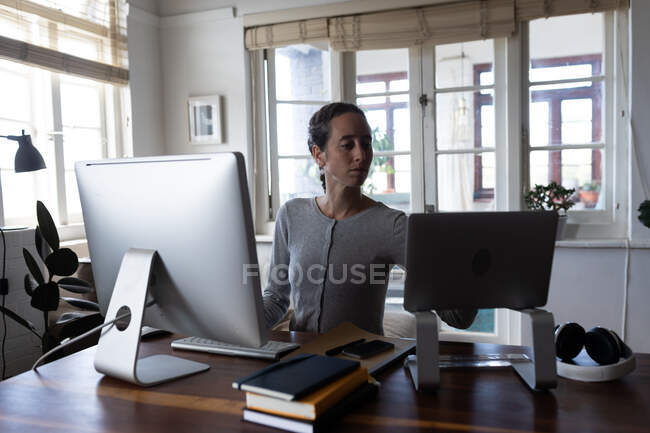 Mulher caucasiana passando tempo em casa, sentada ao lado de sua mesa e trabalhando usando seus computadores desktop e laptop. Distanciamento social e auto-isolamento em quarentena . — Fotografia de Stock
