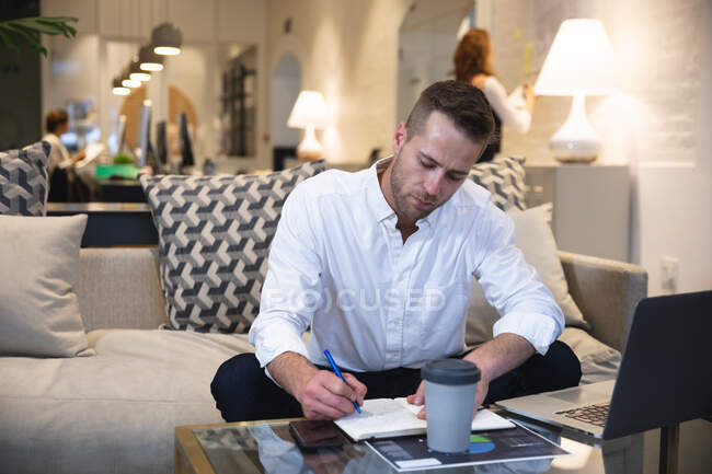 Hombre caucásico creativo de negocios trabajando en una oficina moderna informal, sentado en un sofá y tomando notas, con colegas trabajando en el fondo - foto de stock