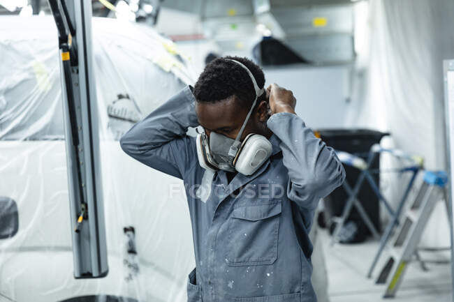 Mecánico masculino afroamericano que usa overoles trabajando en un taller de autos del municipio, poniéndose su máscara respiratoria antes de pintar un auto. - foto de stock