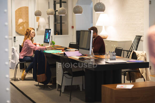Creativos de negocios caucásicos femeninos y masculinos que trabajan en una oficina moderna informal, sentados en escritorios y usando computadoras, tomando notas - foto de stock