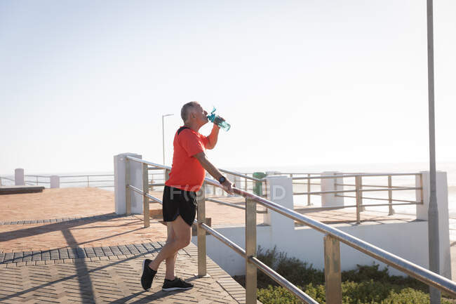 Vista lateral de un hombre mayor caucásico maduro haciendo ejercicio en un paseo marítimo en un día soleado con cielo azul, tomando un descanso, de pie, bebiendo agua - foto de stock