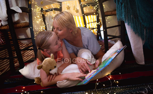 Vue de face d'une femme caucasienne profitant du temps en famille avec sa fille à la maison ensemble, couchée dans une tente dans le salon, lisant un livre, embrassant et souriant sa fille, avec sa fille embrassant son ours en peluche — Photo de stock