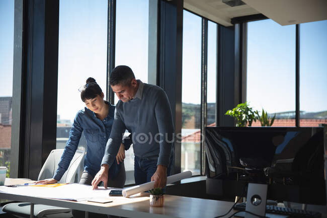 Une femme d'affaires asiatique et un homme d'affaires caucasien travaillant dans un bureau moderne, debout près d'un bureau, regardant des plans et parlant, avec leurs collègues travaillant en arrière-plan — Photo de stock