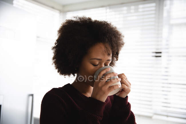 Вид спереду змішаної раси жінка розслабляється вдома, насолоджуючись випивкою чашки кави, тримаючи її обома руками, з закритими очима — стокове фото
