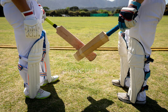 Vista laterale sezione bassa di due giocatori di cricket maschi che indossano bianchi e pastiglie di cricket, in piedi sul campo e attraversando i loro pipistrelli da cricket — Foto stock