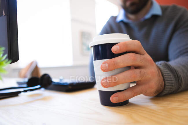 Крупный план кавказца, работающего в современном офисе, сидящего за столом и держащего кофе на вынос — стоковое фото